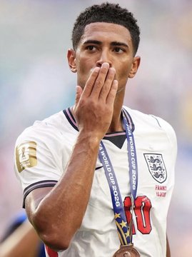 A photo of soccer player Jude Bellingham wearing an England national team jersey with a bronze FIFA World Cup medal around his neck, blowing a kiss.
