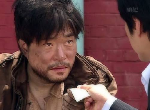 A still from the Korean drama featuring a weathered, teary-eyed man in a worn jacket looking intently at another person holding a small white object.