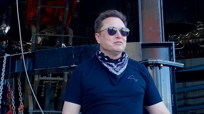 Elon Musk wearing sunglasses and a black bandana around his neck, standing in what appears to be an industrial facility or rocket manufacturing site.