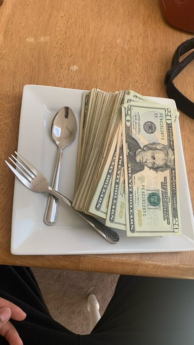 A thick stack of US twenty-dollar bills sits on a white square plate next to a fork and spoon, with a person's legs visible beneath a wooden table.