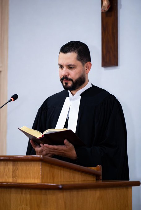 A bearded man in a black judicial robe with white preaching bands reads from a Bible while standing at a wooden podium with a microphone.
