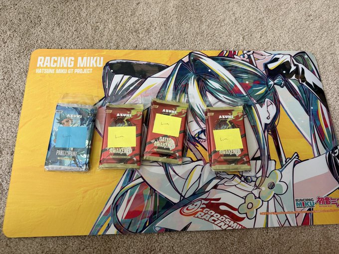 A 'Racing Miku' playmat featuring anime artwork, with four sealed booster packs (labeled 'Participant' and 'Awakened') laid on top of it.