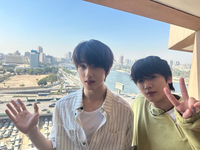Two young men posing on a balcony with a cityscape and waterway behind them; the man on the left waves while the man on the right makes a peace sign.