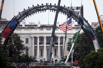 The image shows a large white archway with an American flag flying above it, possibly at the White House. The archway appears to be under construction or being prepared for an event. In the background, there are several people gathered around the site, likely observing and assisting in the process. A crane is also present near the center of the image, indicating ongoing work on the structure.