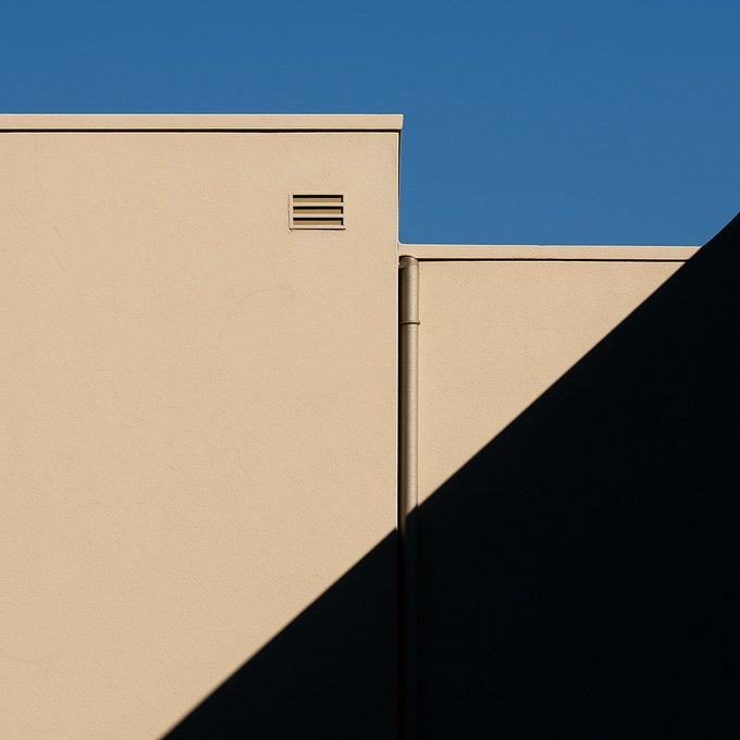 A minimalist architectural photograph of a beige building wall with a small vent, a downspout, and a strong diagonal shadow against a clear blue sky.