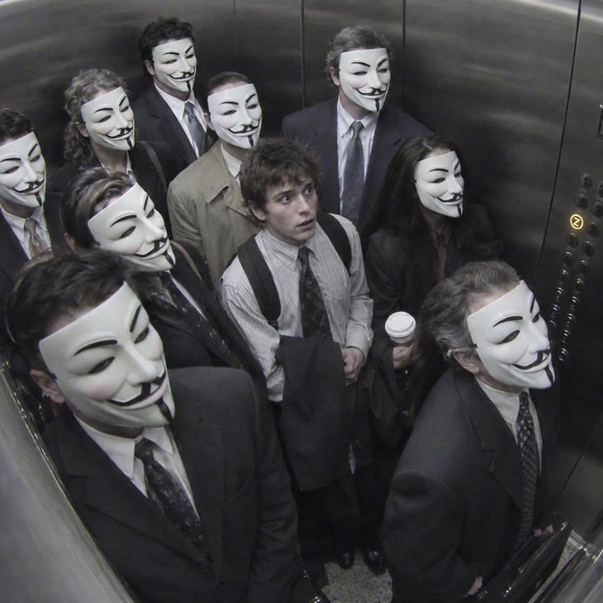 A photograph of a crowded elevator where most people wear Guy Fawkes masks while one unmasked man in the center stands out, holding a coffee cup.