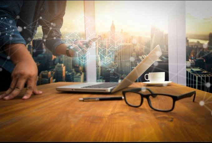 A stock photo showing a person's hands resting on a wooden desk next to an open laptop, glasses, a pen, and a coffee cup, with a city skyline and network graphics overlaid in the background.