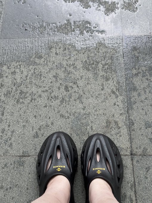 A person wearing black "BINANCE" branded slip-on clogs stands on a wet stone-tiled pavement, likely after rain. The image complements the post by showcasing one of Binance's branded merchandise items, echoing the author's mention of receiving Binance peripherals as gifts.