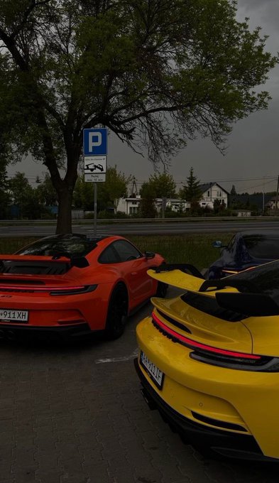 Two sports cars, a yellow Porsche and a red Porsche with Ukrainian license plates, are parked in a lot under a cloudy sky near a parking sign.