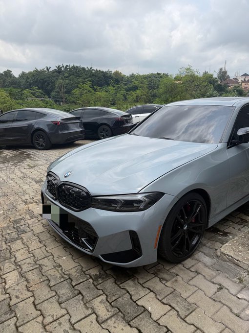 A light gray BMW sedan parked on a paved surface with other cars and greenery in the background; the license plate is blurred.