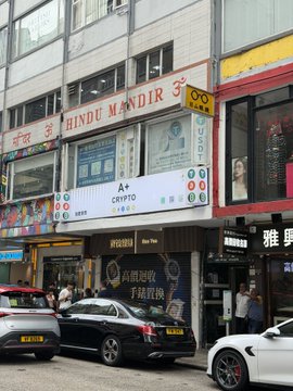 A street view of a Hong Kong storefront displaying signs for "Hindu Mandir," "A+ Crypto," "99 Exchange," and "Mybint." The image relates to the post about exchanging currency (otc) in Hong Kong without KYC, illustrating the local over-the-counter money exchange options available in the area.
