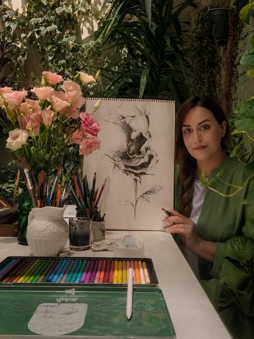 A woman in a green sweater sits at an art table surrounded by flowers and plants, posing with her pencil sketch of a rose displayed on an easel behind her colorful pastel set.