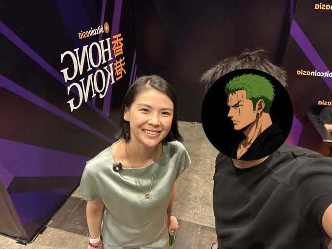 A smiling woman in a gray shirt poses next to a person whose face is obscured by a One Piece anime character (Zoro) sticker, standing in front of a "Bitget x Hong Kong" branded backdrop at a promotional event.