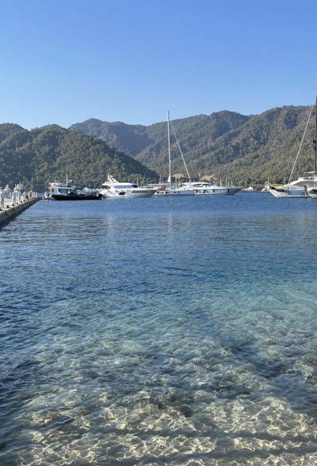 Crystal-clear turquoise water gently laps at the shore of a tranquil marina, where several white yachts and sailboats are docked against a backdrop of lush, forested mountains under a bright blue sky. The serene coastal scene perfectly complements the "Gm my friends, Happy weekend! 🌊" greeting, evoking a peaceful, leisurely vibe ideal for a relaxing weekend by the sea.