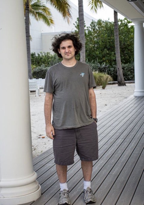A man with curly hair wearing a gray t-shirt and khaki shorts stands on a wooden deck between white pillars, with palm trees and a sandy area visible behind him.