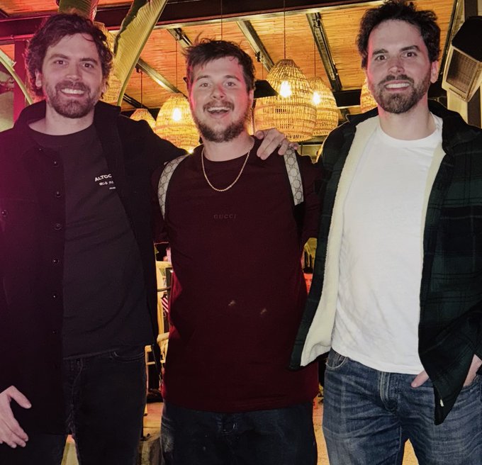 Three friends pose together in a warmly lit restaurant with wicker pendant lamps overhead, including one wearing a maroon "GUCCI" t-shirt in the center, celebrating the Altcoin Daily team's 2 million follower milestone on X in January. The image visually backs the caption's bullish message about crypto community growth, as the same crew has since grown to 2.5M followers.