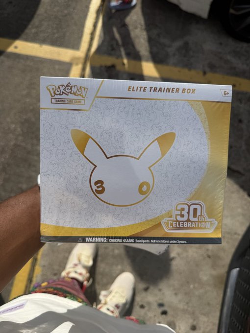 A first-person view of someone holding a sealed Pokémon Trading Card Game 'Elite Trainer Box' featuring a 30th Celebration design with a gold Pichu silhouette.