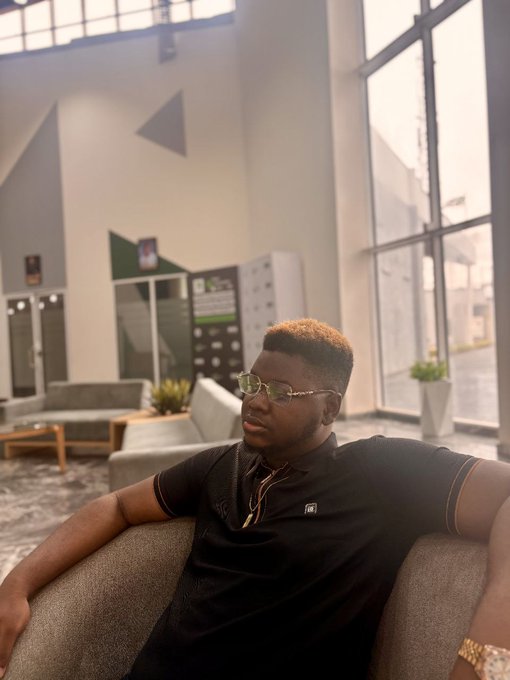 A man wearing glasses, a black polo shirt, a gold chain, and a gold watch lounges confidently in a modern, sunlit lobby with geometric wall designs and large windows. The image complements the post by presenting "bee" as polished and approachable, visually reinforcing his professional yet personable brand in crypto marketing.