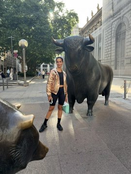 A person posing next to a large bull statue on a city street, holding a small shopping bag.