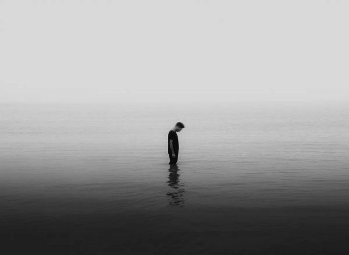 A minimalist black and white photograph of a lone figure standing waist-deep in a calm, misty body of water, with their head bowed down, creating a reflective silhouette.
