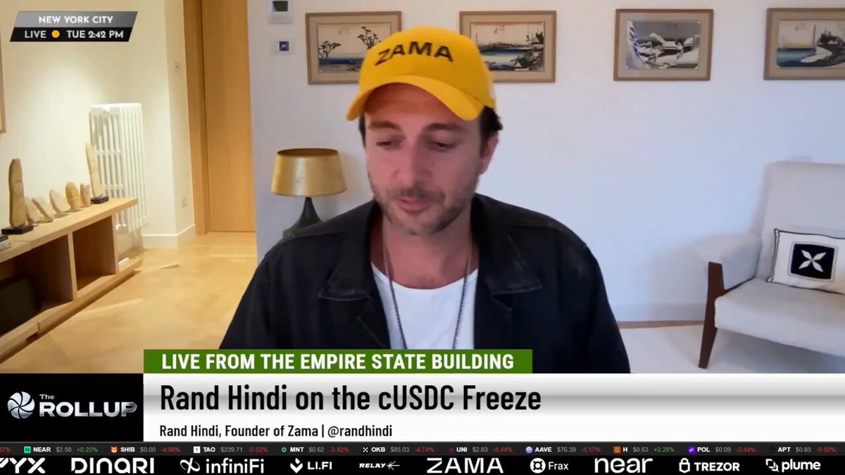 The image shows a man wearing a yellow hat sitting in front of a TV screen displaying a news segment about a partnership between Zama x T-REX RWA, which is an institutional money movement platform. The man appears to be watching or listening intently as the news unfolds on the screen.