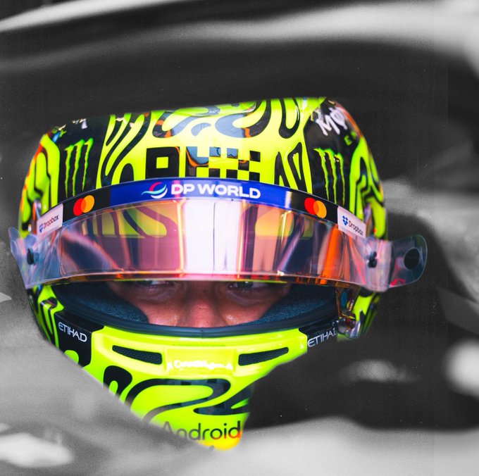 A Formula 1 driver peers through a neon yellow helmet emblazoned with "DP World," "Monster," "Android," and "Etihad" logos, eyes focused intently ahead—capturing the intense concentration required as he prepares to push his limits on the Baku street circuit.