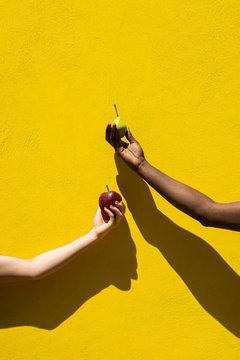 Two hands reach toward each other against a vibrant yellow wall, one holding a green pear and the other a red apple.