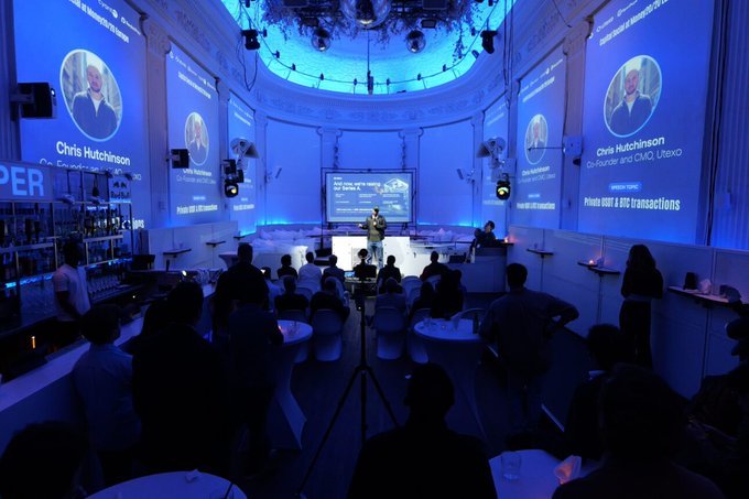 The image shows an event taking place inside a large room with blue lighting, where people are gathered around a stage. A man is standing at the front of the room giving a presentation or speech. The audience consists of several individuals seated at tables and chairs, attentively listening to the speaker. There are also some people standing in the background near the back of the room.

The event appears to be related to Bitcoin or cryptocurrency, as there are banners hanging on the walls that read "Bitcoin", "Cryptocurrency", and "Stablecoins". The audience seems engaged and interested in what the speaker has to say about the topic at hand.
