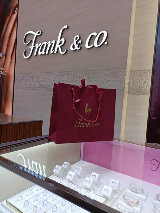 A maroon "Frank & Co." shopping bag sits on a glass jewelry display case showcasing rings and bracelets, with the store's logo mounted on the wall behind it. The image illustrates the post's theme of purchasing gold jewelry as a long-term savings investment, contrasting traditional gold buying with the Bitcoin alternative being considered.
