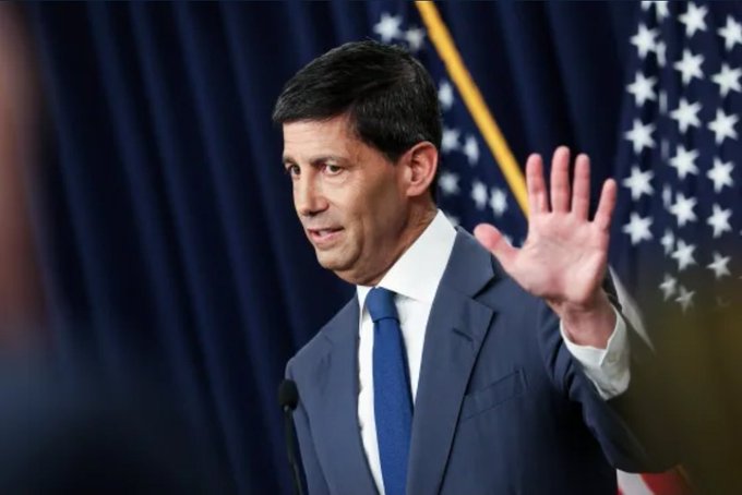 A photograph of a man in a dark blue suit and tie speaking at a podium with American flags visible in the background, appearing to be a government or political figure.