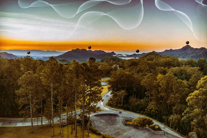 A surreal landscape featuring a sunset over mountains and forested hills with a winding road, overlaid with wavy translucent ribbons in the sky and small floating spheres.