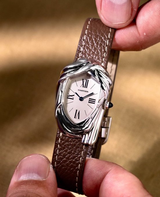 A close-up of a hand holding a silver Cartier watch with a white dial, Roman numerals, and a brown leather strap.