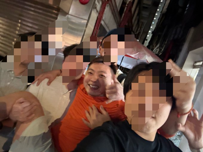 A selfie of a smiling man in an orange shirt being embraced by two people whose faces have been pixelated for privacy.
