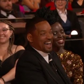 A man in a black suit laughs joyfully alongside a woman in a sparkly gold dress at what appears to be a formal awards event, with other well-dressed attendees visible in the background. The couple's expressions of surprised delight and amusement reflect the post's humor about unexpectedly massive gains exceeding initial expectations, capturing that "I can't believe my luck" feeling of watching a memecoin investment far surpass its original target.