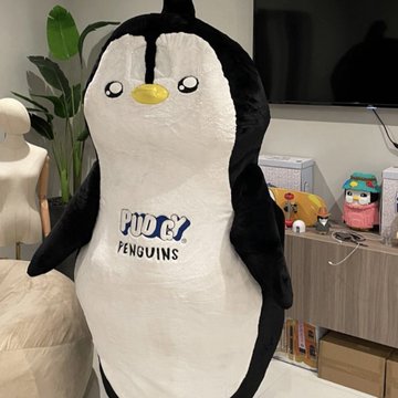 A large plush penguin mascot costume with 'PUGG PENGUINS' written on its white belly stands indoors near a plant and a TV.