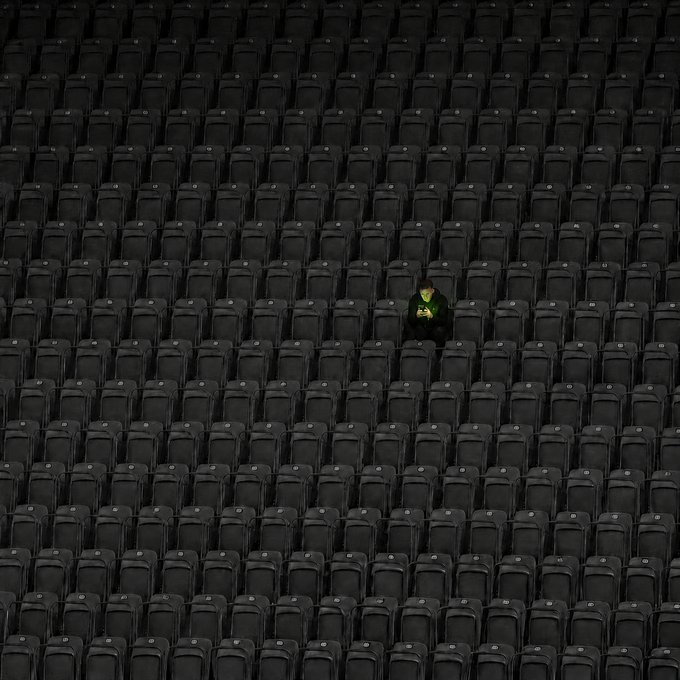 A lone spectator sits in a vast expanse of empty black stadium seats, illuminated only by the glow of a phone screen. The image powerfully illustrates the caption "The conviction comes before the crowd," depicting a single individual holding firm despite the absence of others around them.