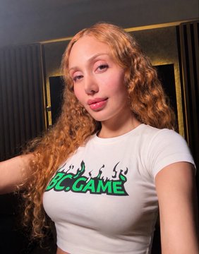 A selfie of a woman with long curly reddish-blonde hair wearing a white t-shirt with green 'DCC.GAME' text, posing in what appears to be an indoor setting.
