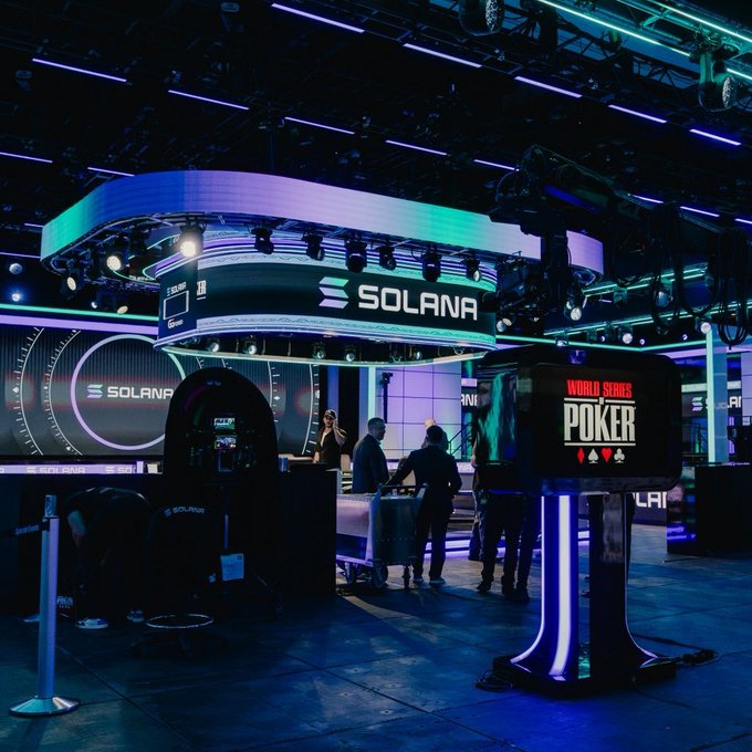 A dimly lit, blue and purple illuminated room featuring a large overhead circular display with the 'SOLANA' logo, alongside a 'WORLD SERIES OF POKER' sign, with several people standing in the space.