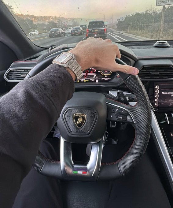 The image shows a person wearing a black sweatshirt, sitting inside a car with their hand on the steering wheel. They are driving down a street filled with cars, including some that appear to be moving at high speeds. The person's watch is visible as well, indicating they might be keeping track of time during their drive or possibly waiting for someone else in the vehicle.