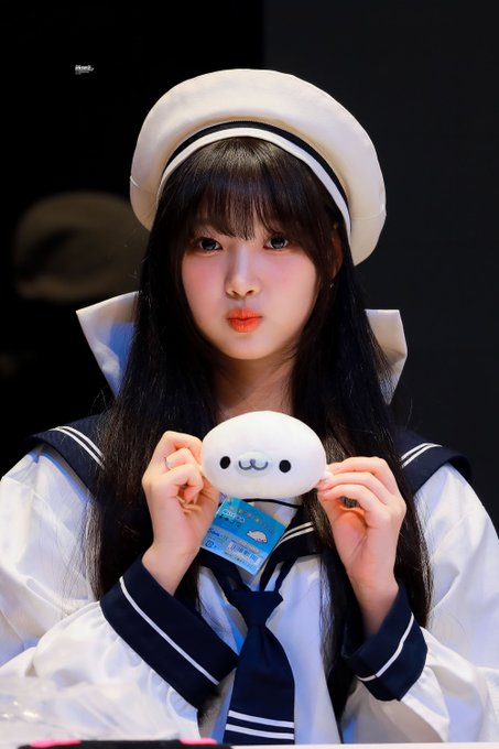 A young woman with long black hair wearing a white sailor hat and matching sailor-style outfit, holding a small white plush toy with a cute face.