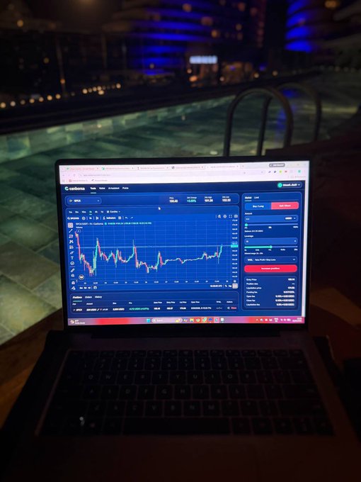 A laptop displays a CoinMarketCap cryptocurrency chart on a balcony at night, overlooking a pool area illuminated with blue lights.