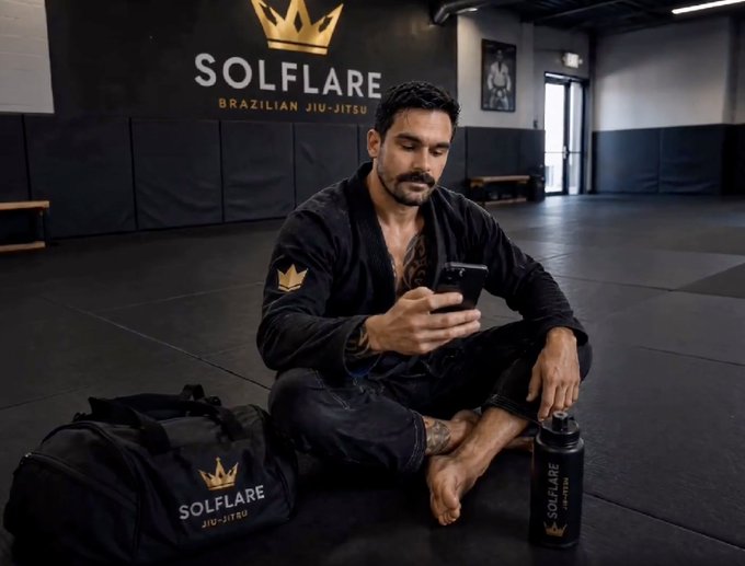 A man in a Solflare Brazilian Jiu-Jitsu gi sits cross-legged on the gym mat, smiling at his phone, with his branded gym bag and water bottle beside him and the "SOLFLARE BRAZILIAN JIU-JITSU" logo on the wall behind him—capturing a relaxed, grateful "gm and happy friday" moment with his team.