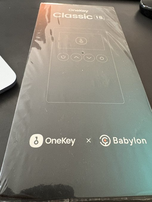 A OneKey Classic 1s hardware wallet box in green packaging featuring a collaboration logo with Babylon, sealed in plastic wrap on a dark surface.