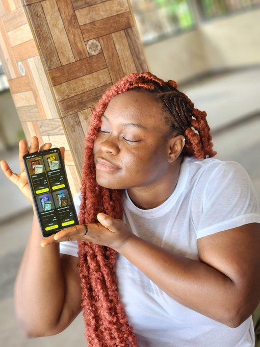 A woman with long red braids smiles peacefully with her eyes closed while holding up a smartphone displaying a colorful app interface.