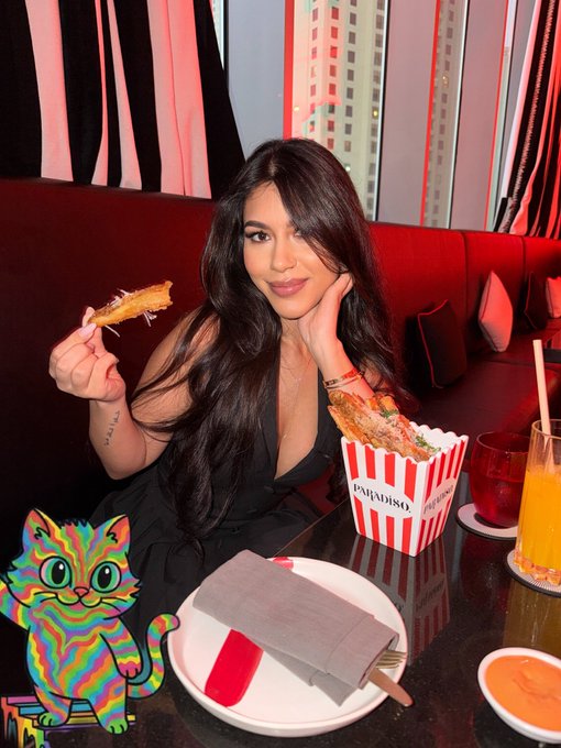 A woman with long dark hair sits at a restaurant table holding a piece of fried food, with a 'PALADISO' popcorn container and a cartoon cat sticker overlay in the corner.