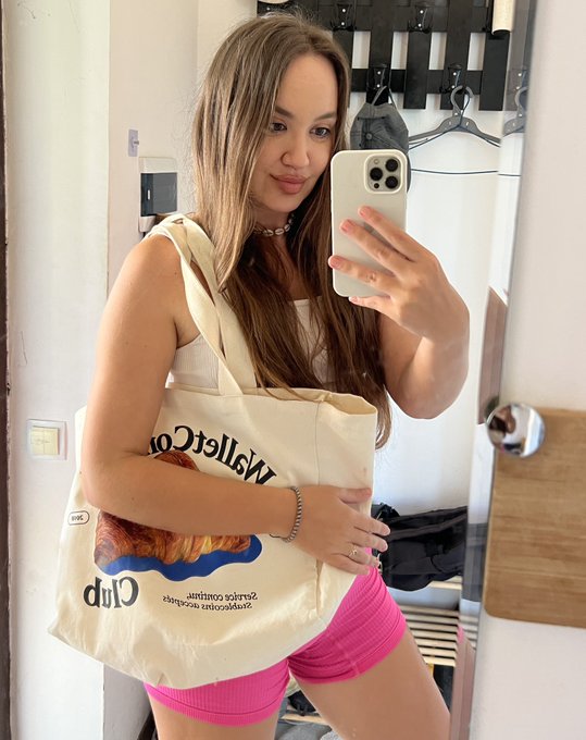 A young woman takes a mirror selfie with a white smartphone, wearing pink shorts and carrying a cream tote bag with 'MeltCo Club' printed on it.
