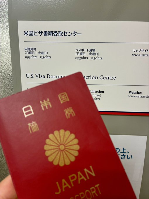A Japanese passport held up in front of a sign for the U.S. Visa Document Collection Centre (米国ビザ書類受取センター), indicating pickup hours of 0300hrs-1330hrs.