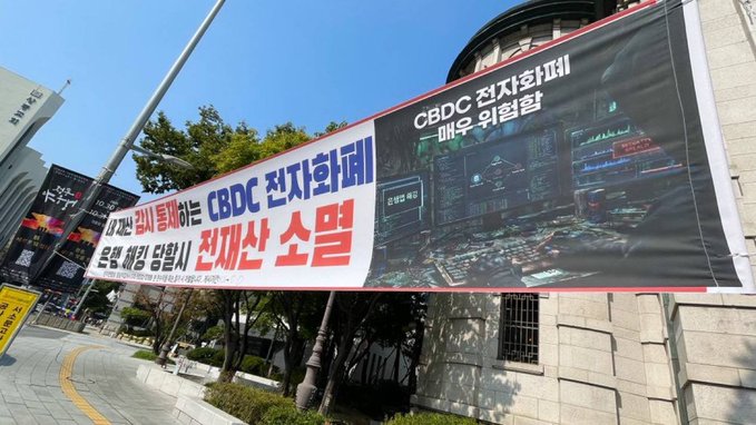 A large outdoor banner reading 'CBDC 전자화폐' and '전재산 소멸' hangs on a street in Korea, warning against electronic currency.