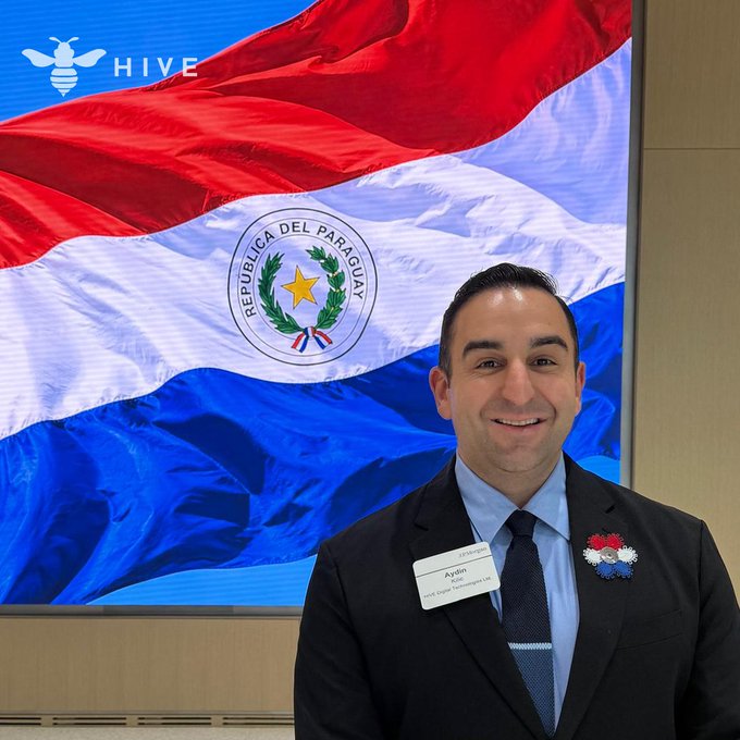 Aydin Kilic, President & CEO of HIVE (visible by the bee logo and his name badge), stands smiling in front of a large screen displaying the flag of Paraguay ("República del Paraguay") at the Paraguay Investment Forum in NYC, illustrating the company's engagement with the country as an emerging innovation hub in Latin America.