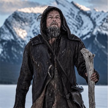 A bearded man in tattered, dark hooded clothing stands in a snowy mountain landscape holding a large wooden staff.